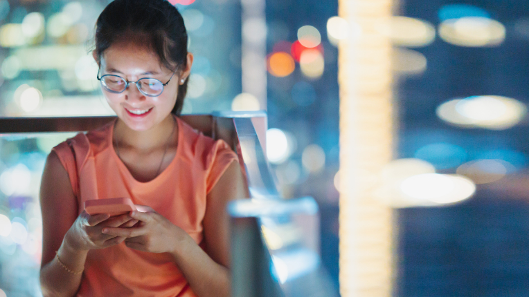 Woman standing on rooftop patio and looking at smart phone amidst urban city lights.